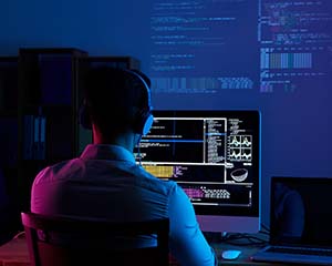 A man sitting in a dark room in front of a computer which is currently displaying code.